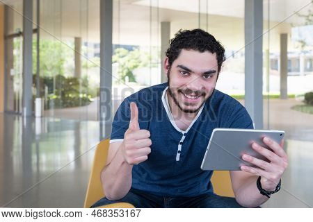 Smiling Young Man Looking At Camera, Making Success Sign With Thumb, Sitting In Lobby Of Office Buil