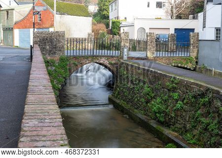 Long Exposure Of A Watefall Flowing Under A Bridge On The River Lim Walkway At Lyme Regis In Dorset.