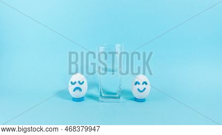 Next To The Half-filled Glass, Two Eggs With A Cheerful And Sad Expression. Concept Of Optimist And