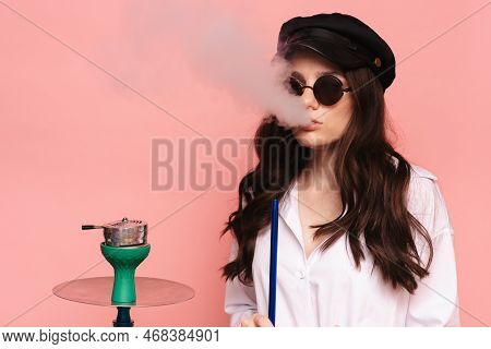 An Attractive Woman In A Cap And Glasses Smokes A Hookah, Sheesha And Enjoys Smoking. Pink Solid Col
