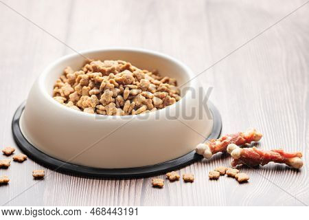 A Bowl Of Dog Food And Treats In The Form Of Bones On A Wooden Floor. Dry Food In Granules.