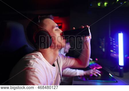 Young Guy Gamer In Headphones Plays A Computer And Drinks An Energy Drink, A Man Sits At A Computer