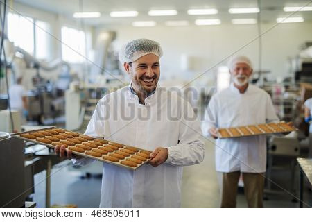 Picture Of Two Happy Smiling Male Employees In Sterile Clothes Carrying Trays Full With Fresh Cookie