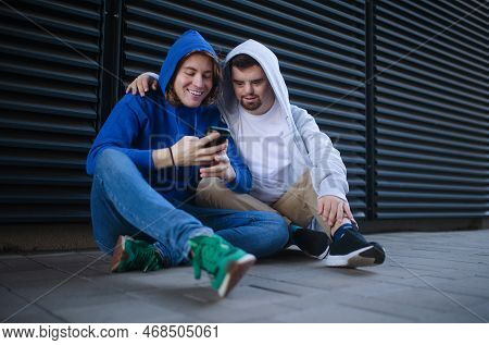 Man With Down Syndrome Resting Outdoor With His Friend, Scrolling Smartphone. Concept Of Friendship 