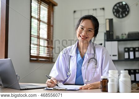 Beautiful Asian Female Doctor In Uniform Sits At Her Desk, Smiling And Looking At The Camera.
