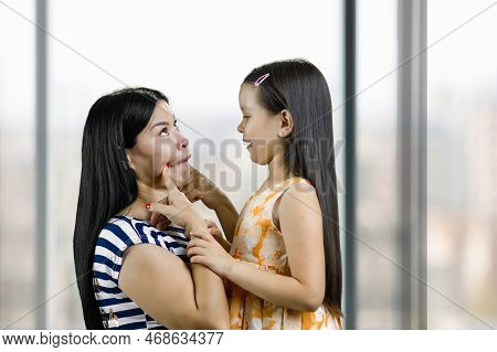 Mother Making Funny Face To Her Little Daughter. Woman Pulling Her Cheeks. Blurred Indoor Windows Ba
