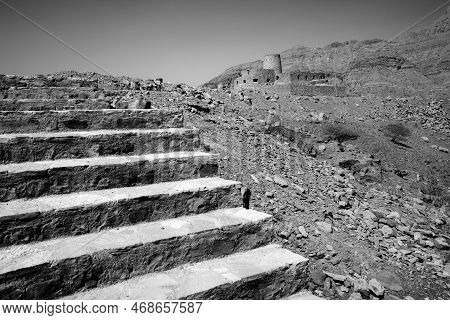 Black And White Shot Of Stone Walls Of Small Medieval Arabian Fort Under Tall Mountain Cliffs. Fortr