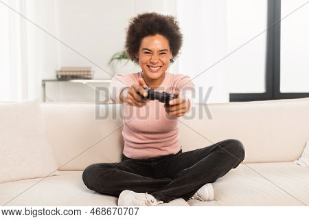 Cheerful Excited Young Mixed Race Woman Sitting On Sofa With Joystick, Playing Computer Game In Livi