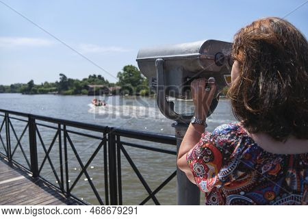 Latin Mature Woman Viewing Through Public Binoculars On The Lujan River Bank In Tigre, Buenos Aires,