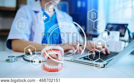 Concentrated Dentist Sitting At Table With Jaw Samples Tooth Model And Working With Tablet And Lapto