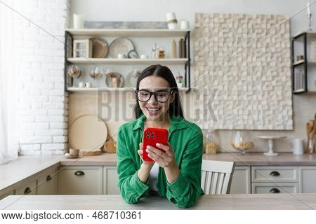 Happy And Smiling Young Teenage Girl Sittiing At Home In The Kitchen And Using The Phone. Receives M
