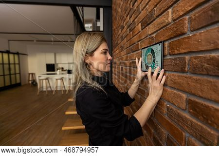 Young Woman Controlling Home With A Digital Touch Screen Panel Installed On The Wall In The Living R