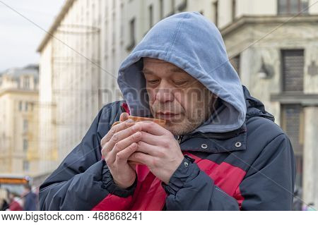 A Homeless Man Of 45-50 Years Old In A Jacket And Hood Holds A Mug With A Hot Drink In His Hands, Cl