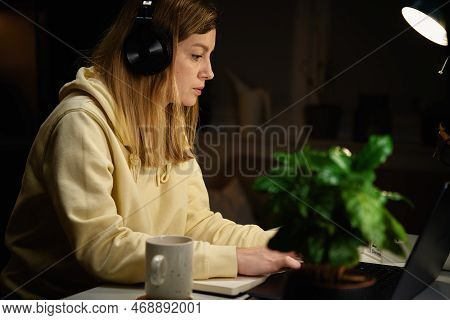 Female Sudent Wearing Headphones Studying At Night Room, Using Laptop. Online Education At Home.