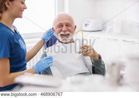 Senior Man Sitting At Dentist Chair Holding Saliva Ejector While Doctor Fixing His Tooth