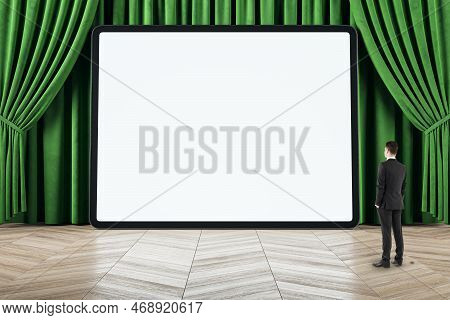 Businessman Looking At Empty White Mock Up Tablet, Green Curtains And Wooden Flooring. Cinema, Stage