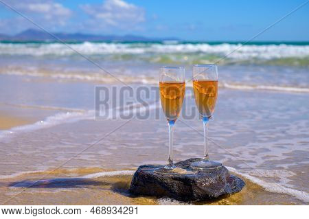 Two Glasses Of Rose Champagne Or Cava Sparkling Wine Served On White Sandy Tropical Beach And Blue O