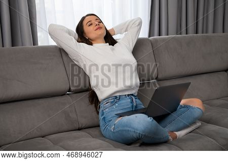 Relaxed Young Woman Lounges On A Comfortable Sofa, Arms Crossed Behind Her Head And Holding Laptop O