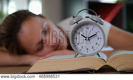 Tired Woman Sleeps On Table With Book And Alarm Clock. Overwork And Study At Night Concept.