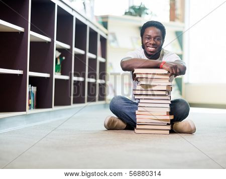 Full length portrait of happy African American student leaning on stacked books while sitting on floor at library