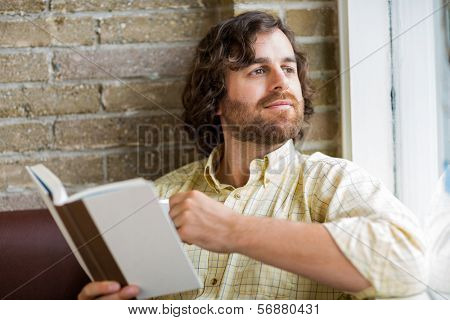 Thoughtful mid adult man with book looking through window in coffeeshop