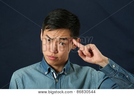 Funny young Asian man pointing his index finger against his temple as crazy sign