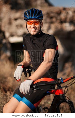 closeup portrait of professional smiling cyclist with his bicycle in the countryside. Background with blurred rock. Sportsman in the black sportwear, helmet and sunglasses. Countryside. Detail. Hands and face.