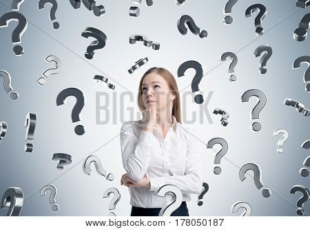 Portrait of a pensive blond girl standing near a gray wall with question marks falling aground her. Concept of too many questions.