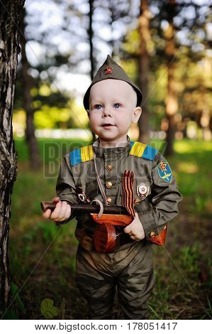 A cute child in the uniform of a Soviet soldier with a St. George ribbon with a toy gun on the background of nature. May 9, February 23, defender, against fascism.