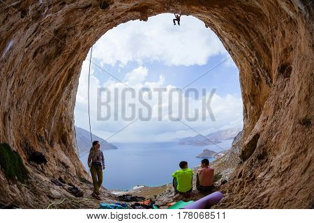 Group of rock climbers in cave: two men resting and talking young male climbing on ceiling of cave his female partner watching him through belay glasses