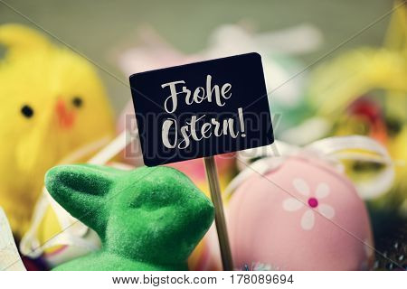 a chalkboard with the text frohe ostern, happy easter in german written in it and a pile of decorated easter eggs of different colors, a green rabbit and a yellow teddy chick in the background