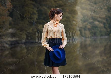 outdoor fashion portrait of sensual stylish girl in vintage clothes with lake and forest on the background. Young woman with romantic hairstyle and floppy hat walks in the forest. Female retro look