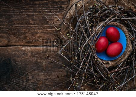 Three Easter red eggs on a blue plate in the nest with spring branches on an old wooden table