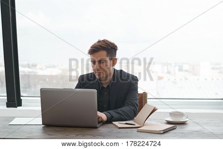 Concentrated man typing online message in blog in office with glass window. Spend working time for social network on the job. Business man using laptop computer with high speed internet for surfing
