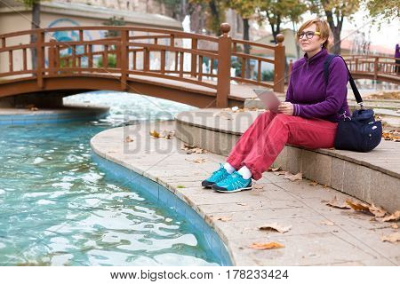 Inspired Woman in casual and sporty Clothing sitting in Park next to Pond and using tablet pc device