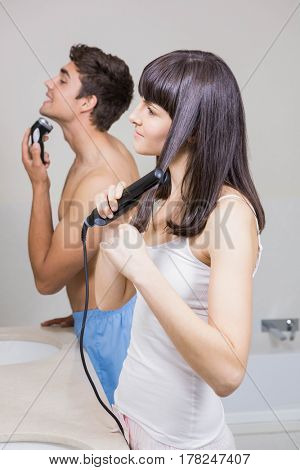 Young man shaving and young woman straightening hair in the bathroom