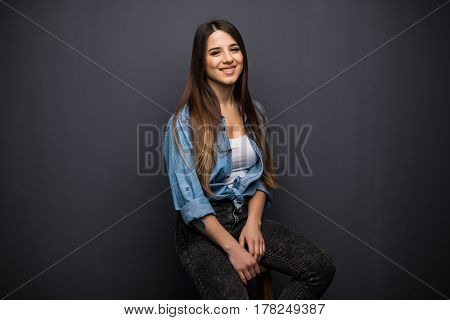 Portrait Of Pretty Young Woman With Long Hair In Casual Shirt Sitting On Stool Chair On Black Backgr