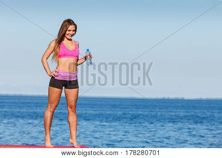 Woman in sportswear takes a break to rehydrate drinking water from plastic bottle resting after sport workout outdoor by seaside