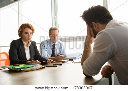 Business representatives of huge company taking interview in one young man. Handsome man sitting in front having stress white interview. Job placement concept.