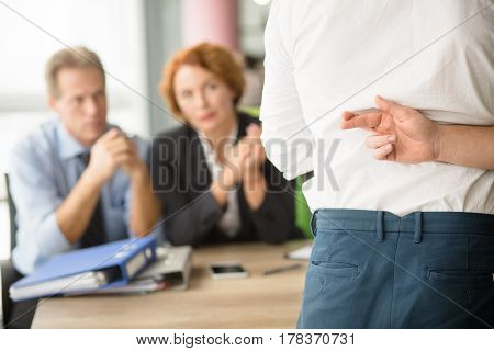 Closeup of young man crossed his fingers before taking interview in office while employees ready to communicate with new applicant.