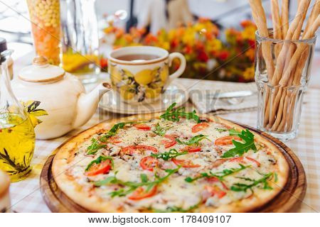 Pizza with cheese, tomato and meat, with cup of tee on served table with plaid tablecloth