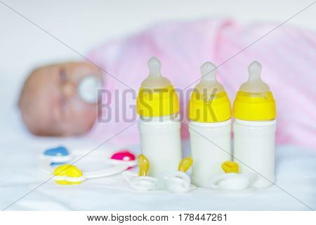 Cute newborn baby girl with nursing bottles, rattle and pacifier. Formula drink for babies. New born child, little girl laying in bed. Family, new life, childhood, beginning, bottle-feeding concept.
