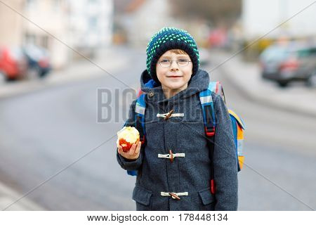 little kid boy with eye glasses on the way to the school and eating fresh apple. child on a street. preschooler wearing warm clothes, glasses and backpack. happy student or people. education concept