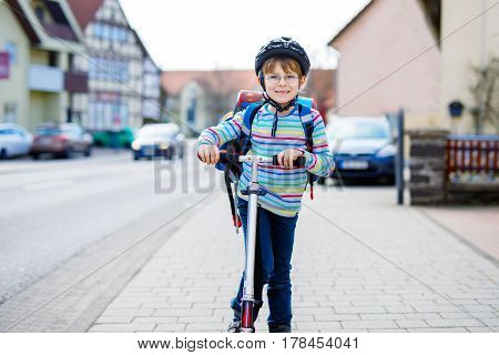 active school kid boy in safety helmet riding with his scooter in the city with backpack on sunny day. happy child in colorful clothes biking on way to school. safe way for kids outdoors to school.