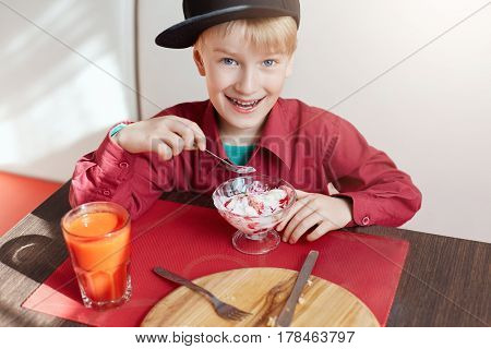 A happy young boy with blue eyes and blond hair dressed in red shirt and stylish cap siting at cosy caffe eating delicious ice-cream and drinking tomato juice looking happily into a camera