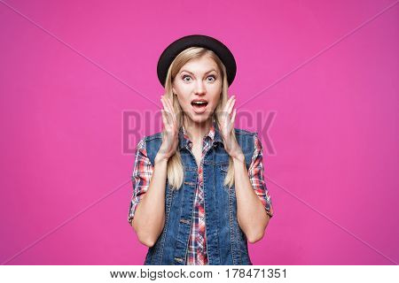 Surprised hipster young woman wearing black hat and denim vest shcoked. isolated on pink stusio background
