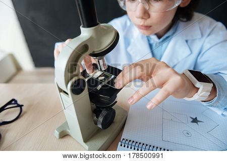 Exploring evolution of microbiology. Skillful gifted capable teenager sitting in the laboratory and enjoying science lesson while makig notes and using microscope