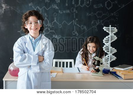 Defending our chemistry project. Smiling delighted skilled boy standing in the lab and expressing positivity while his classmate working on the scince project in the background