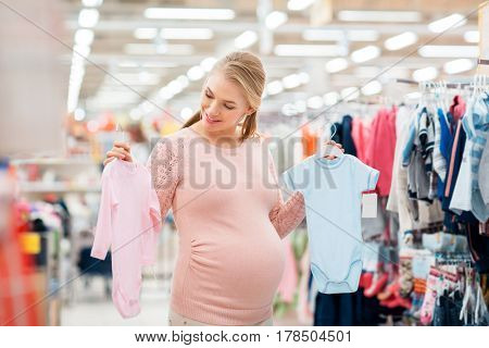 pregnancy, people and shopping concept - happy pregnant woman choosing between blue and pink baby bodysuits at clothing store