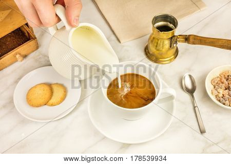 Milk poured into a cup of coffee, with vintage coffee grinder and pot in the background, and cane sugar. Selective focus